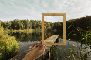 Hand hält Holzrahmen vor Steg an einem See mit Wald im Hintergrund